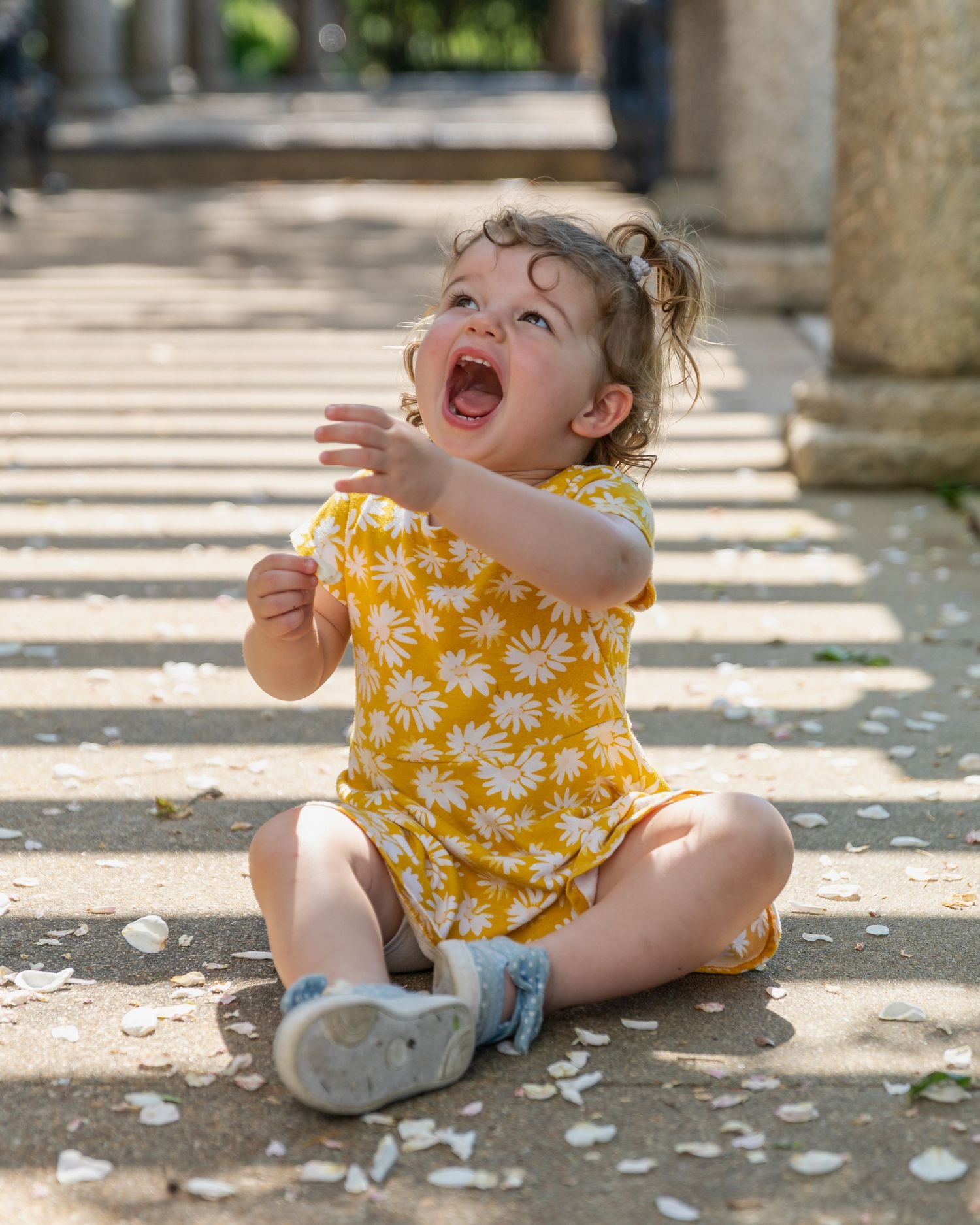 Laughing toddler in yellow floral romper sitting on ground in dappled sunlight.