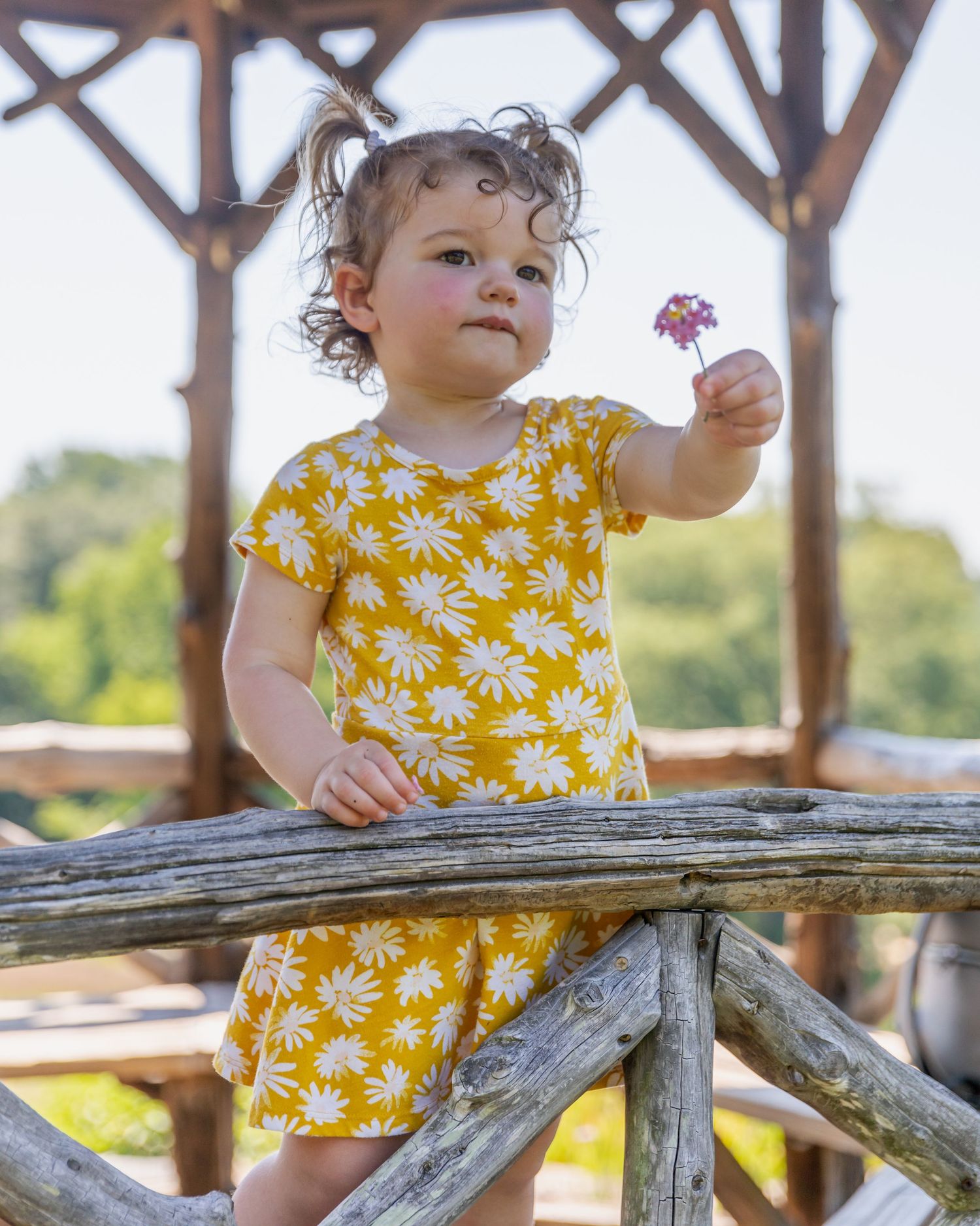 Small child in yellow daisy print dress holding flower while standing at rustic wooden railing.