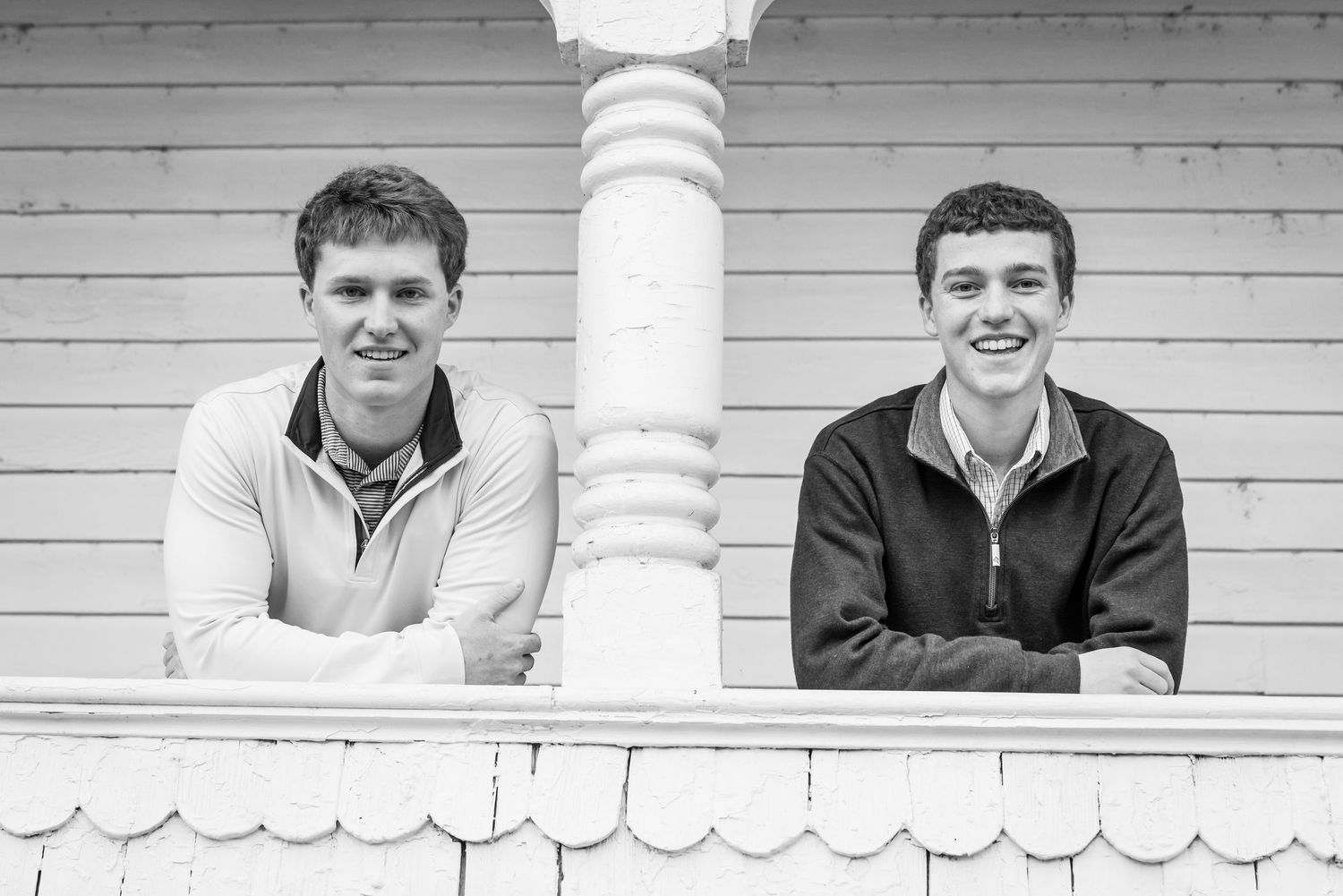 Two young men smiling while leaning on white porch column in black and white photo.