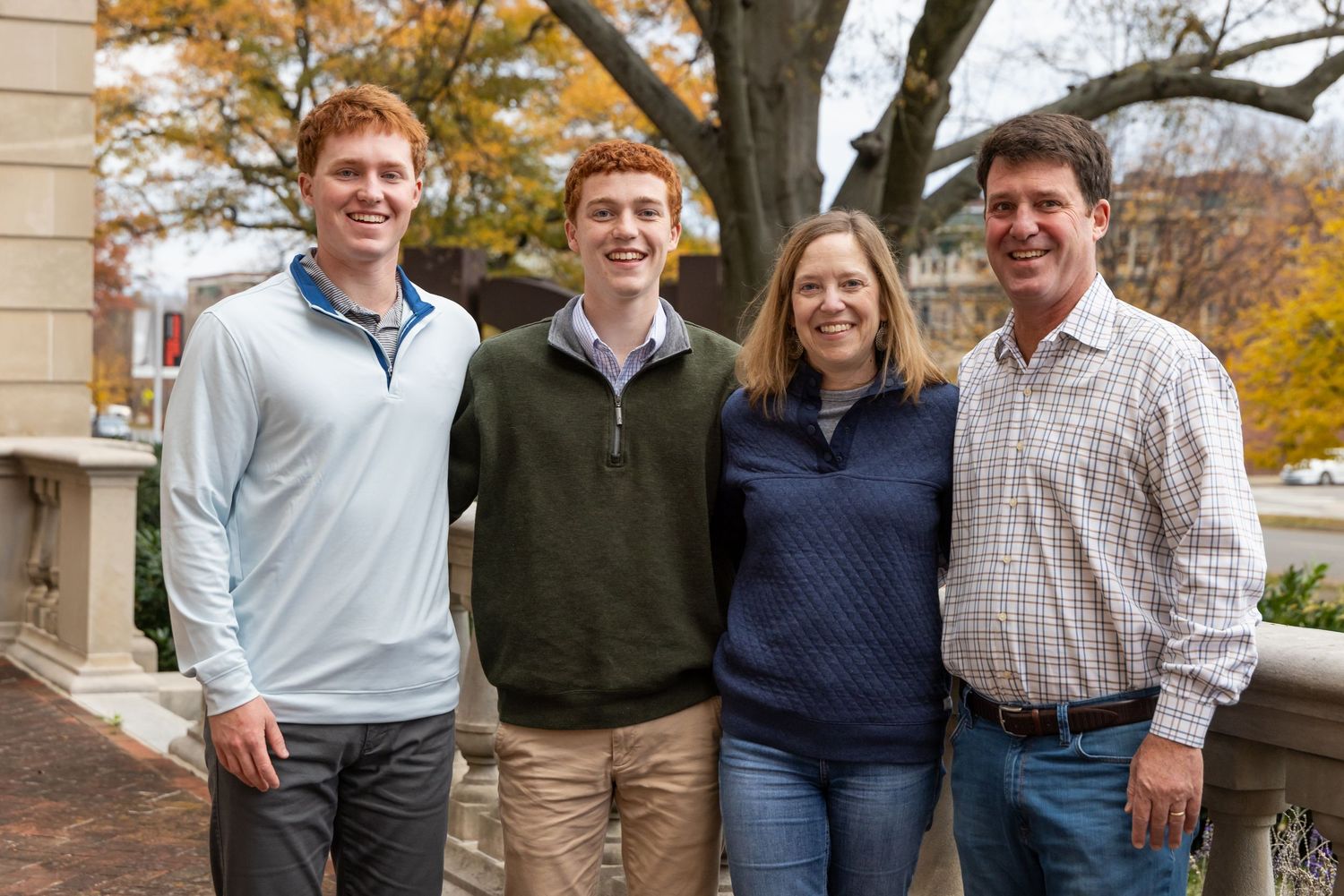 Family of four poses together on autumn day with fall foliage in background.