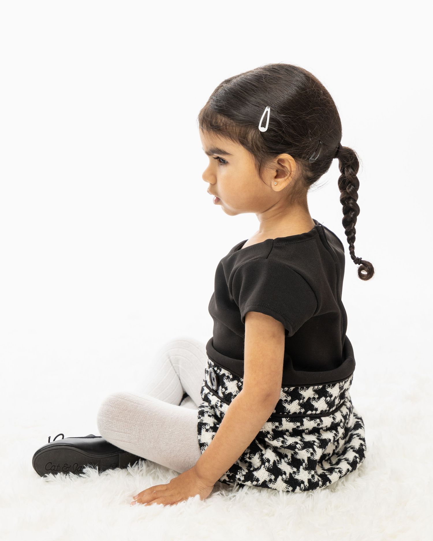 Side profile of young child in black top and houndstooth skirt sitting on white background.