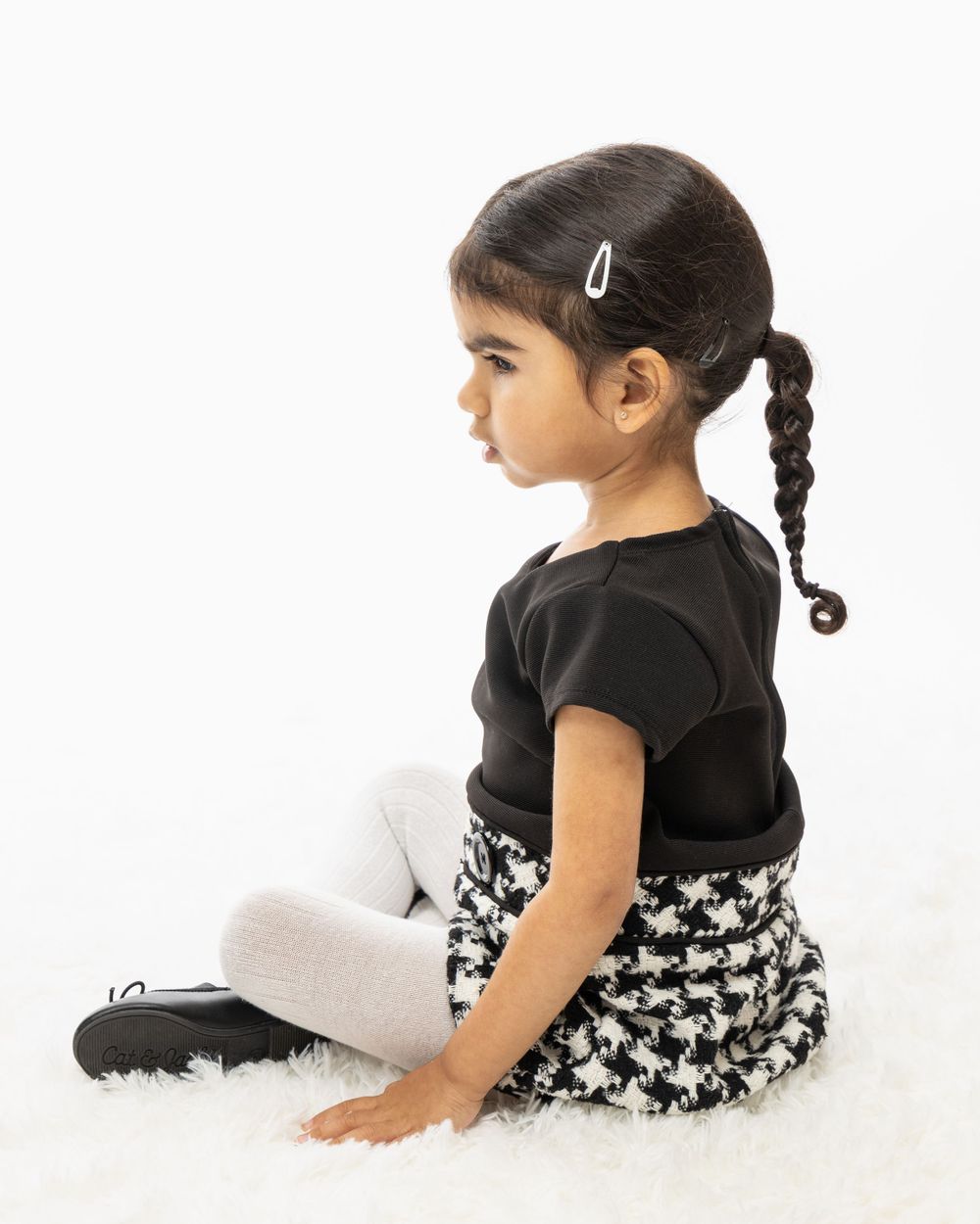 Side profile of young child in black top and houndstooth skirt sitting on white background.
