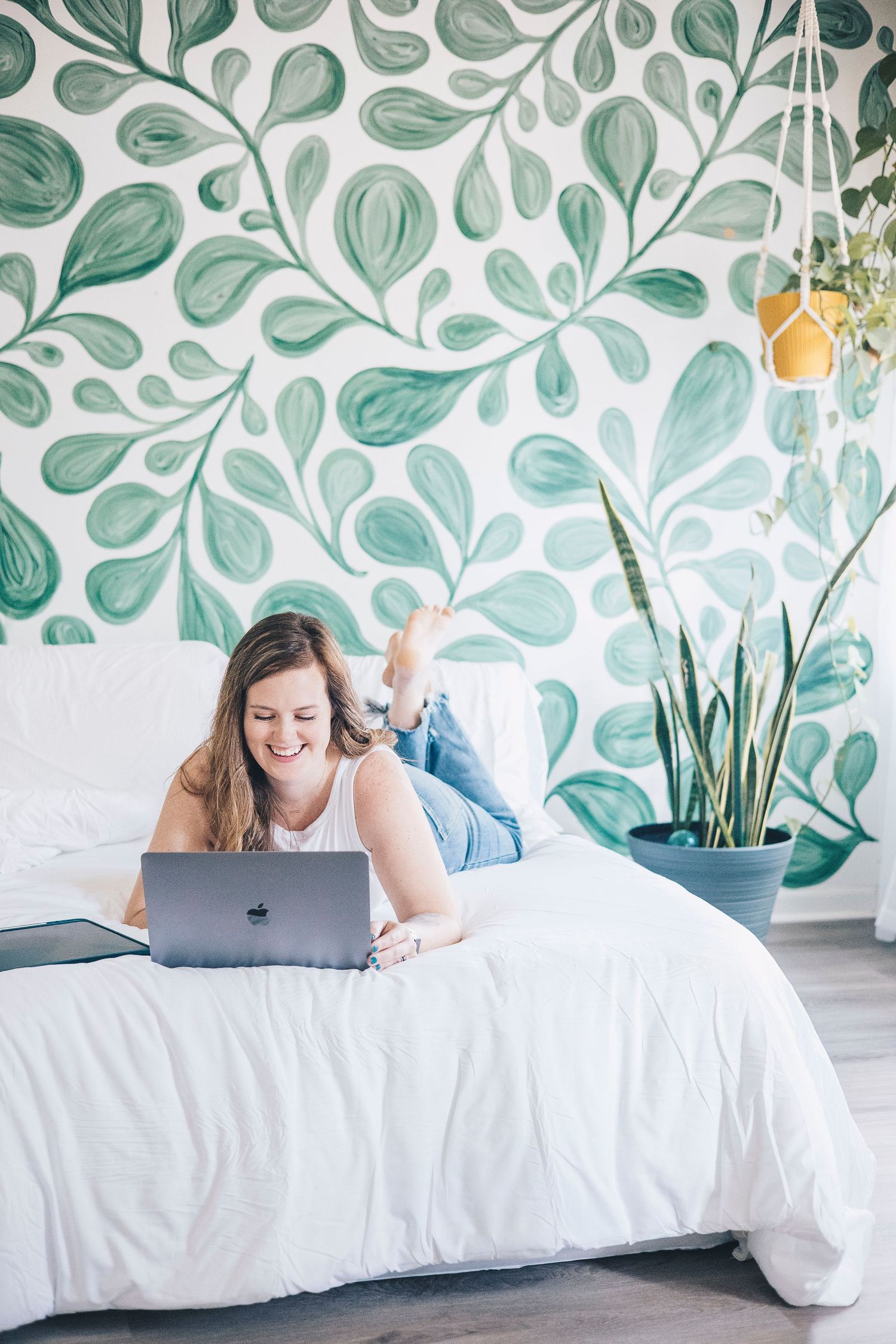 Woman lying on bed with laptop in front of green botanical wall mural.