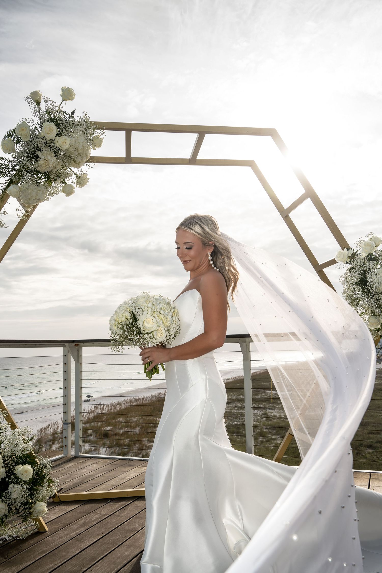 Bride in flowing white dress stands under geometric arch by oceanside.