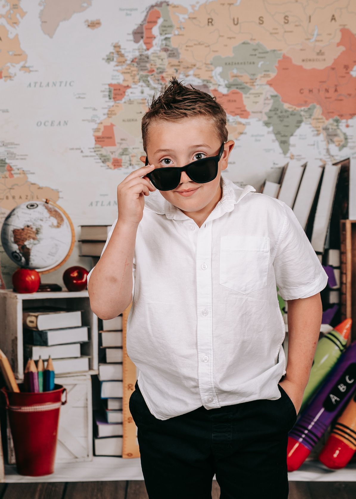 A cool young boy in a white shirt poses with sunglasses in front of a colorful world map backdrop.