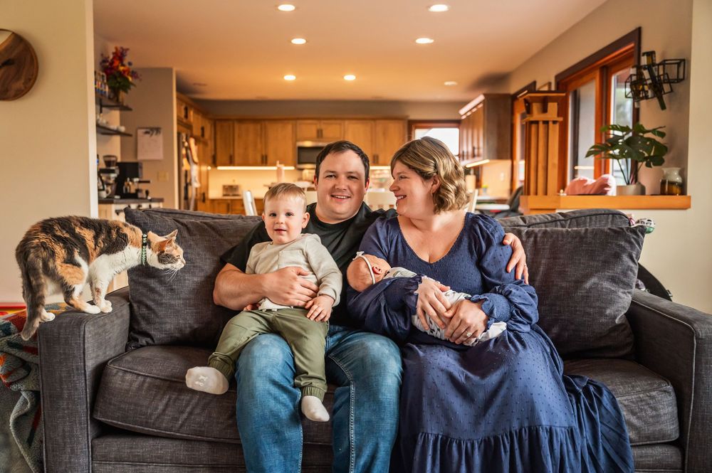 Family members sit together on a gray couch in a cozy living room with warm lighting and wood accents.