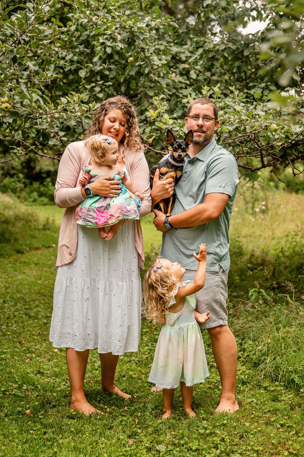 A family shares playful moments together in an apple orchard on a sunny day, wearing casual summer attire.