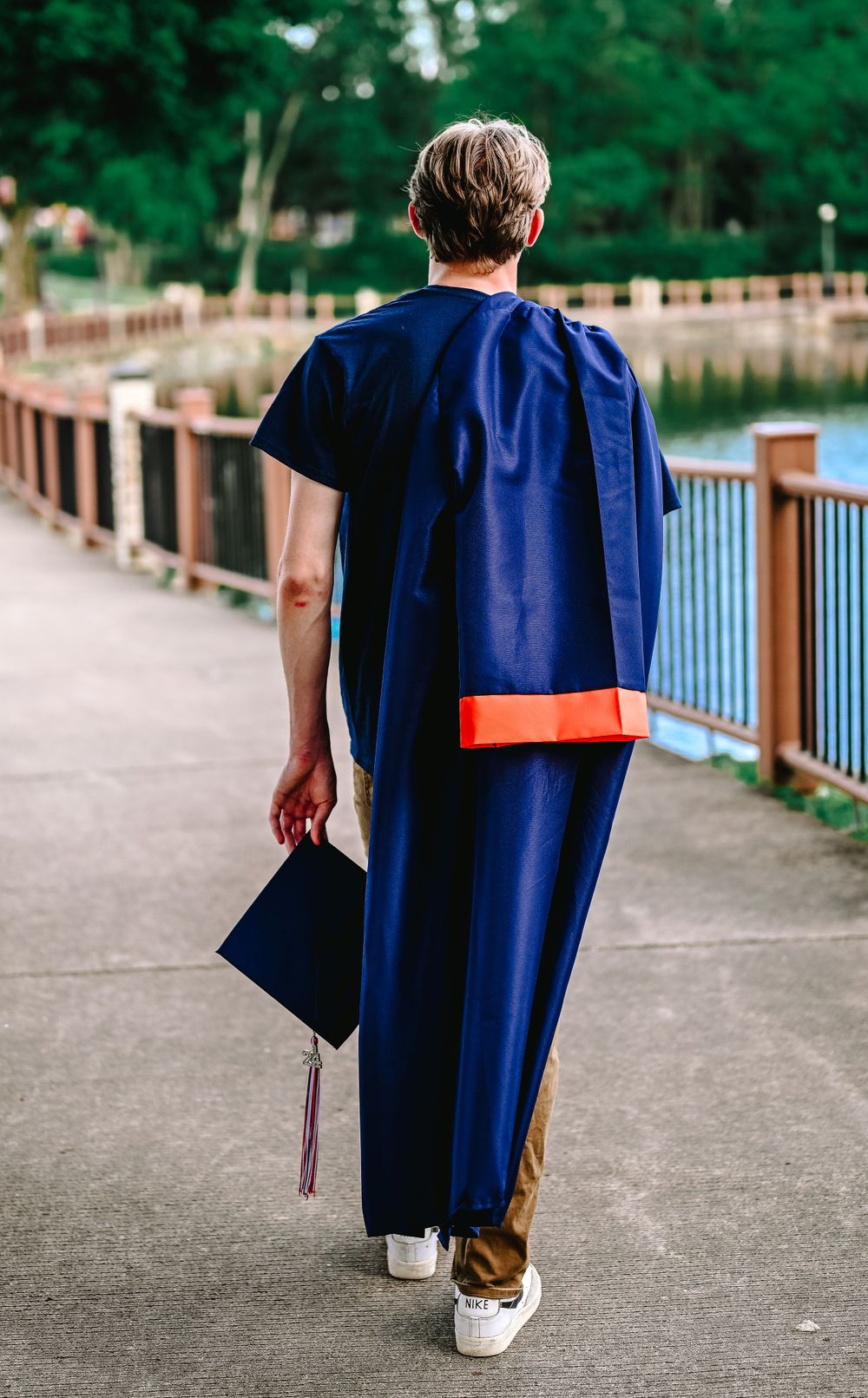 Graduate in blue cap and gown walking on boardwalk path, view from behind.