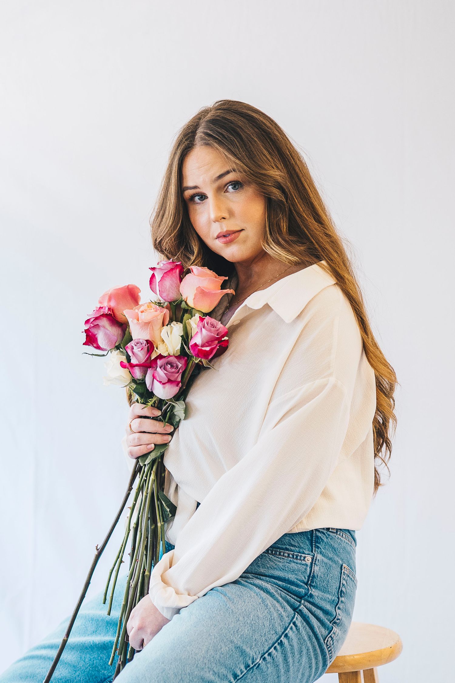 Young woman with long hair holding a bouquet of pink roses, wearing a white blouse and jeans.