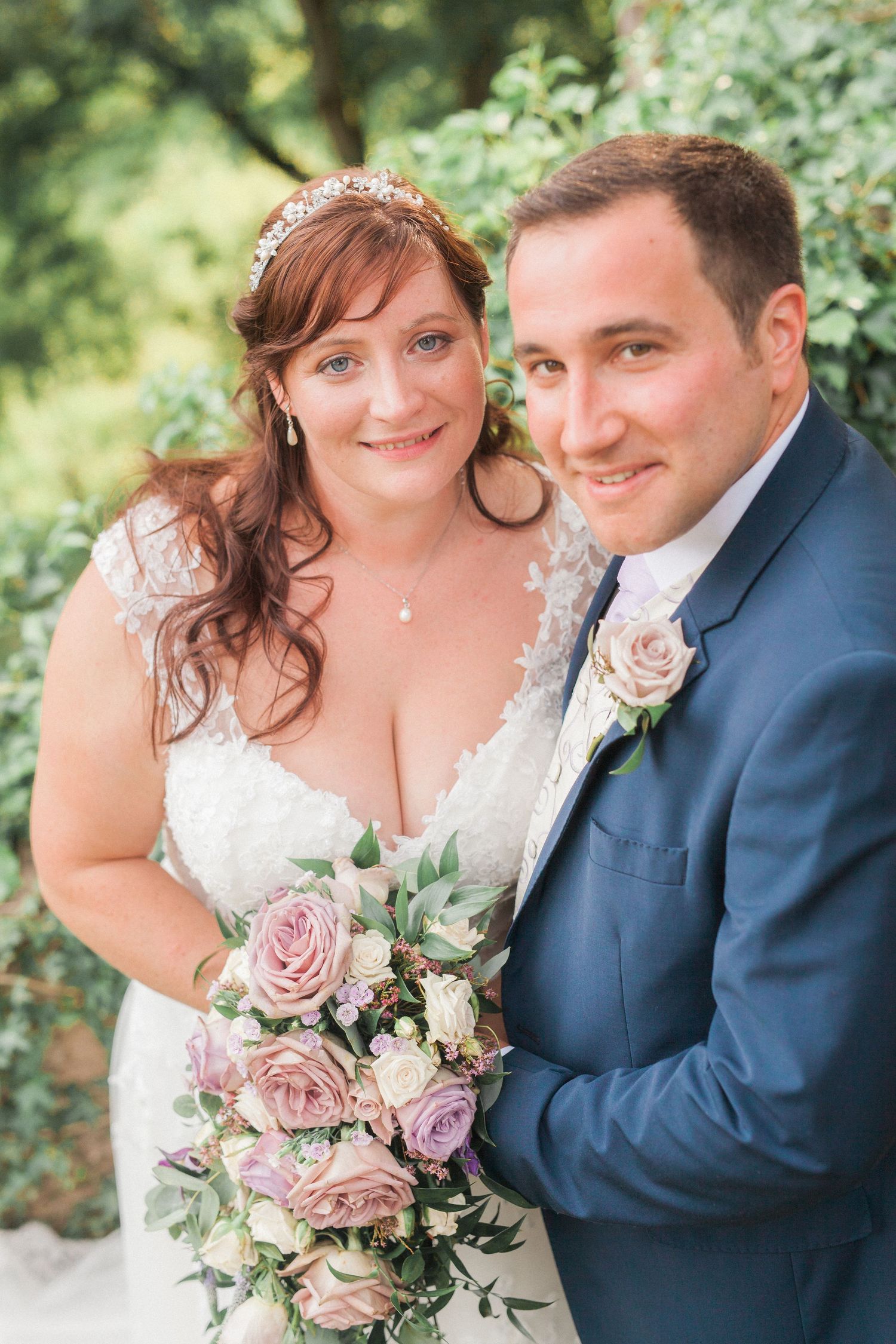 Wedding couple poses together outdoors with pink rose bouquet and formal attire in garden setting.