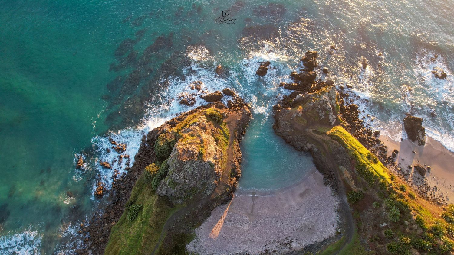 Aerial view of rugged coastal rock formations meeting turquoise ocean waves with a sandy beach cove.