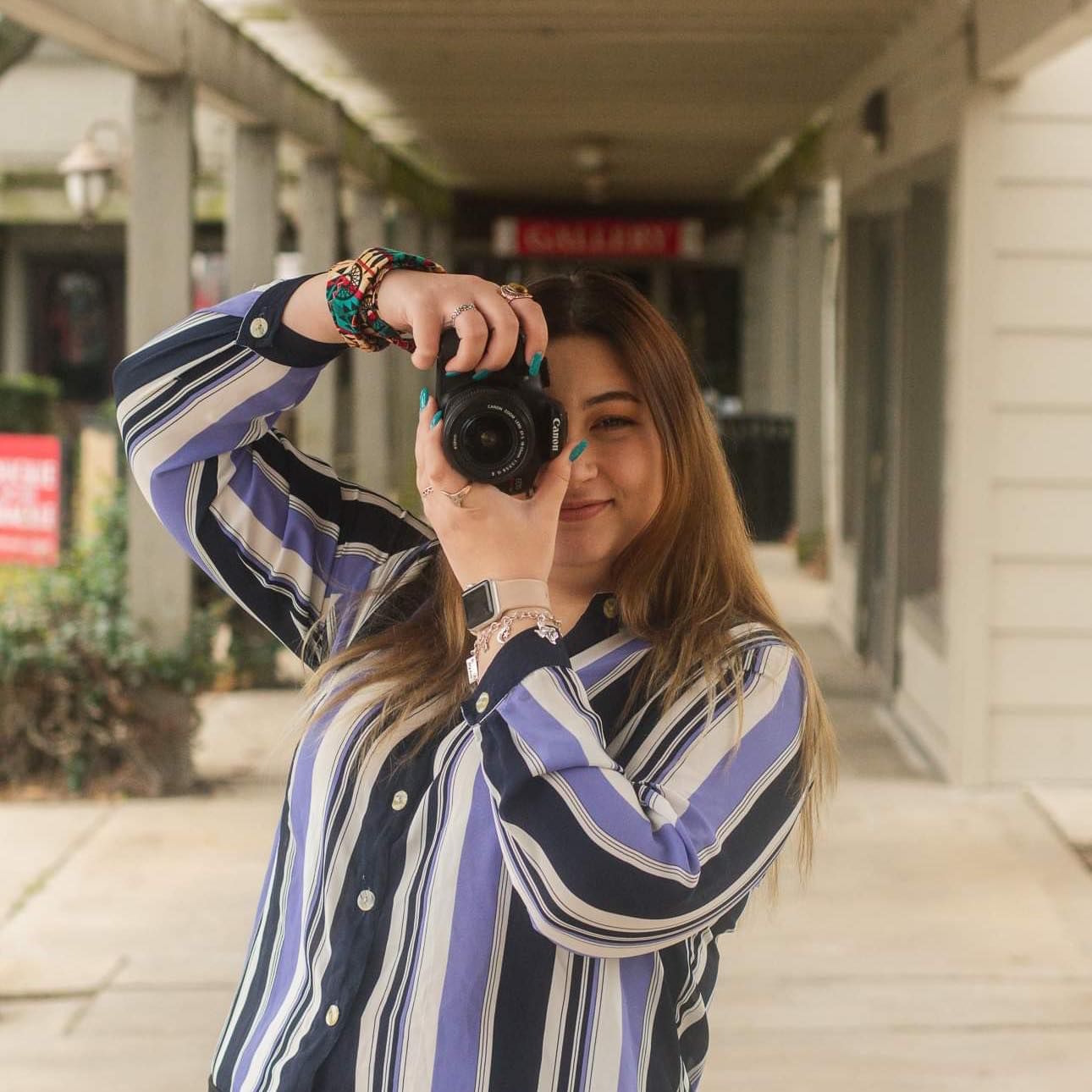 Person wearing a striped blue and white shirt takes a photo with a digital camera in covered outdoor walkway.