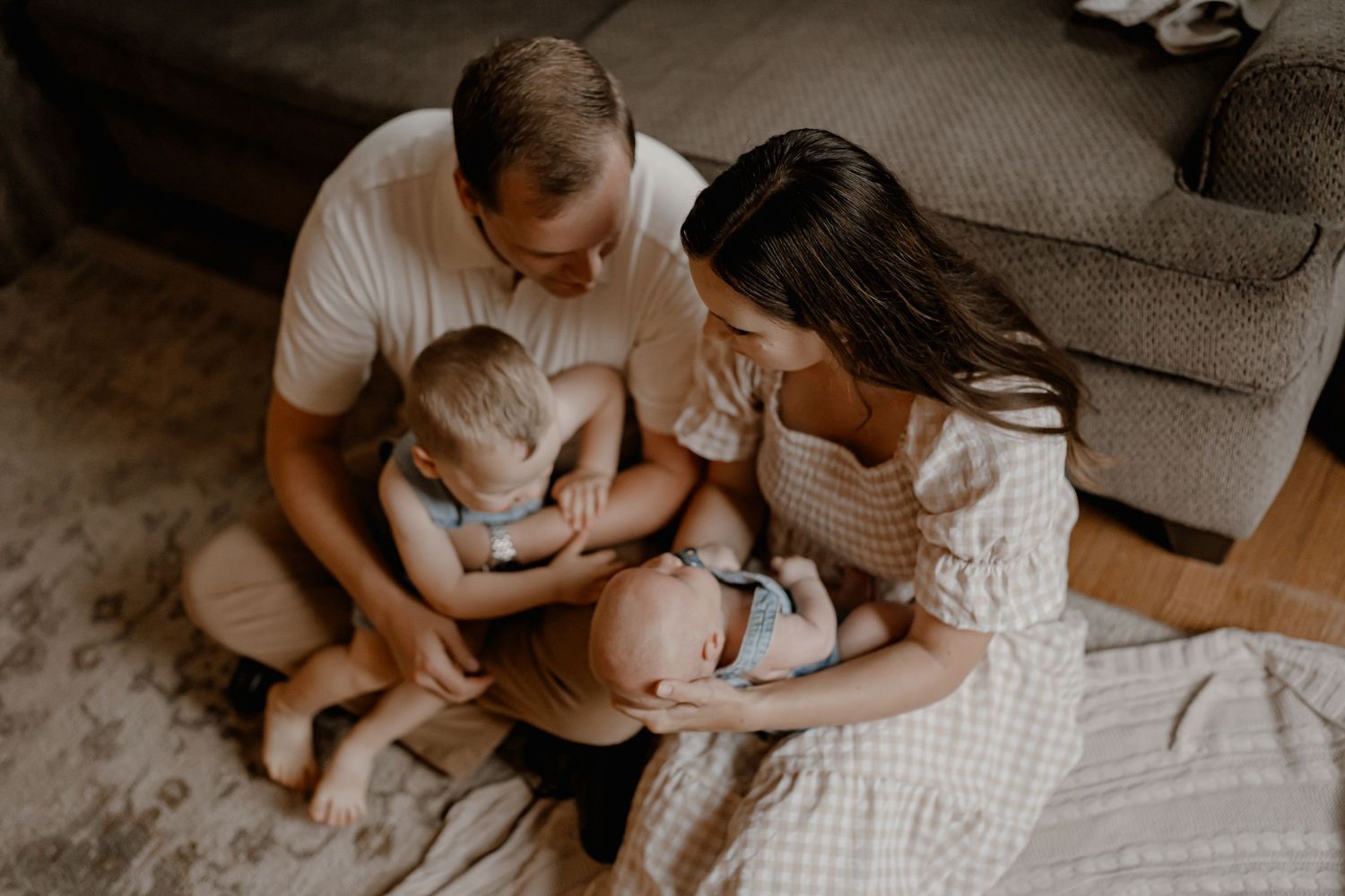 Family sits together on living room floor with pet dog while sharing tender moment in neutral toned setting.