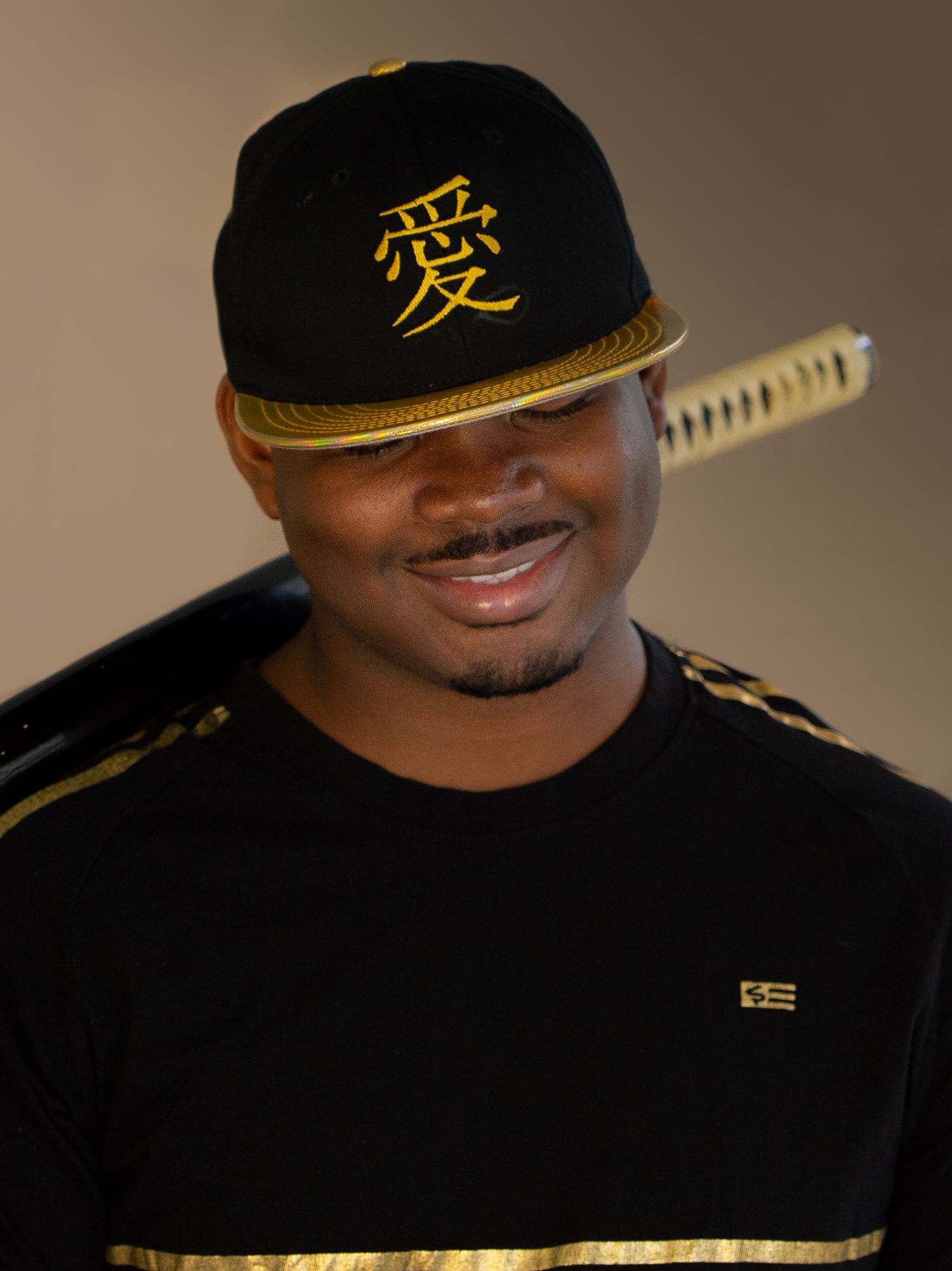 Man smiling wearing black gold snapback hat with Chinese character and katana sword posing confidently.