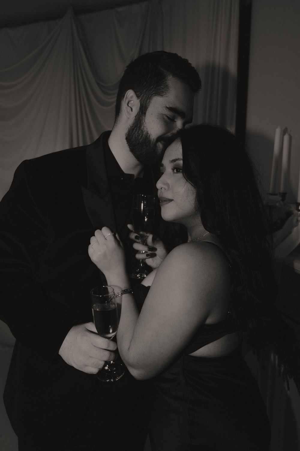 A romantic black and white photo of a couple embracing while holding champagne glasses in a dimly lit room.