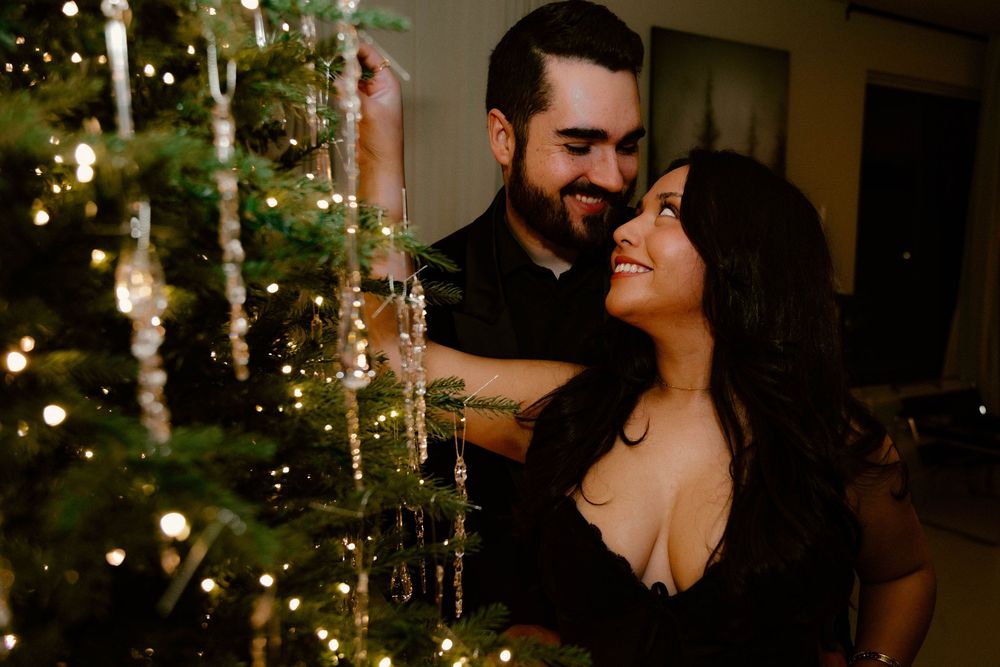 A romantic couple embraces by a glowing Christmas tree in a dimly lit room, creating an intimate holiday moment.