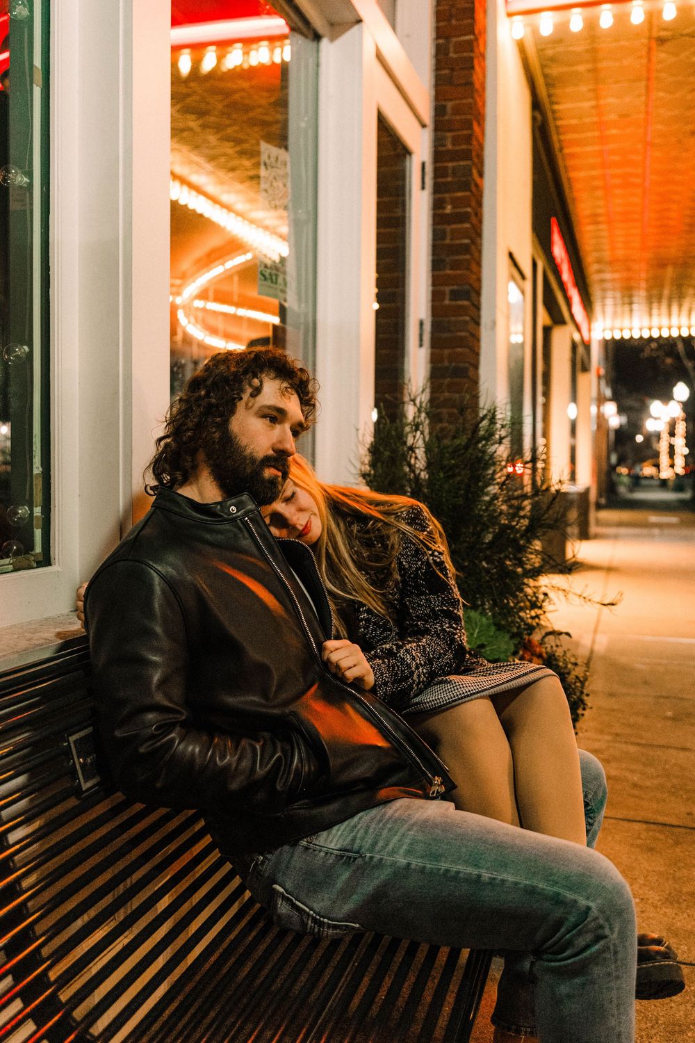 Person in leather jacket sitting on bench outside storefronts with string lights at night.