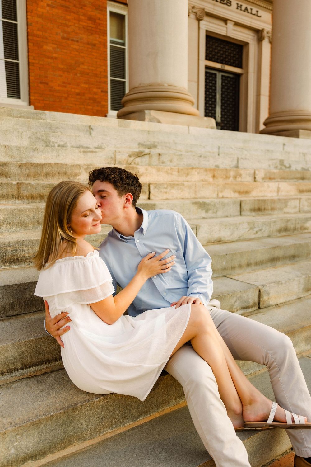 A romantic couple sits on stone steps of a brick building sharing a tender moment in light summer attire.