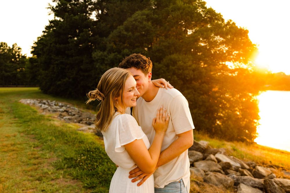 A couple in white outfits embraces on a grassy shoreline during a vibrant golden sunset.