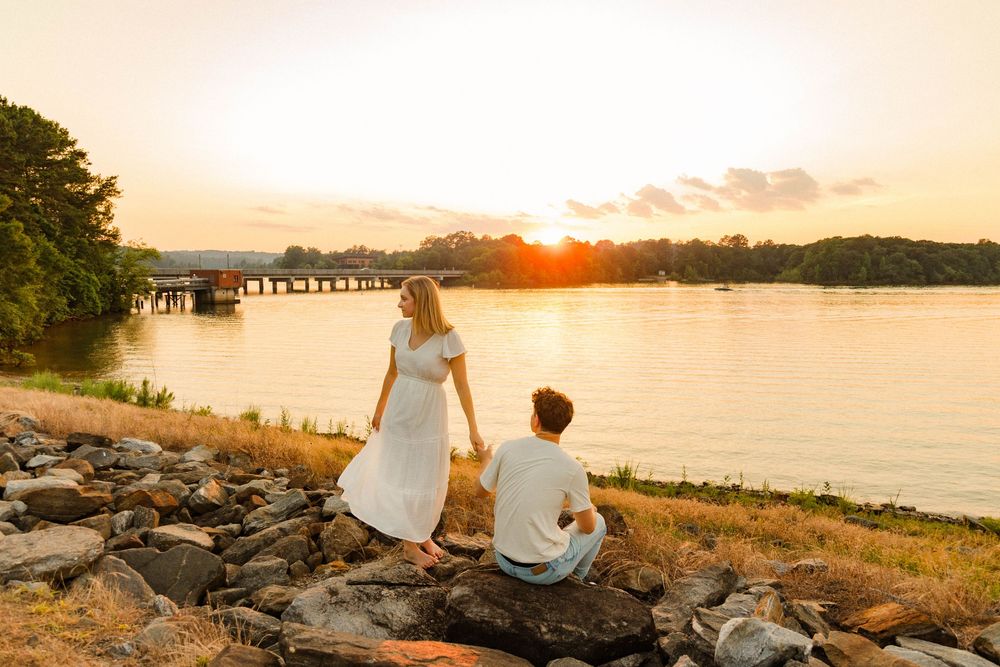Two people sit on rocky shore watching golden sunset over calm lake with wooden pier in distance.