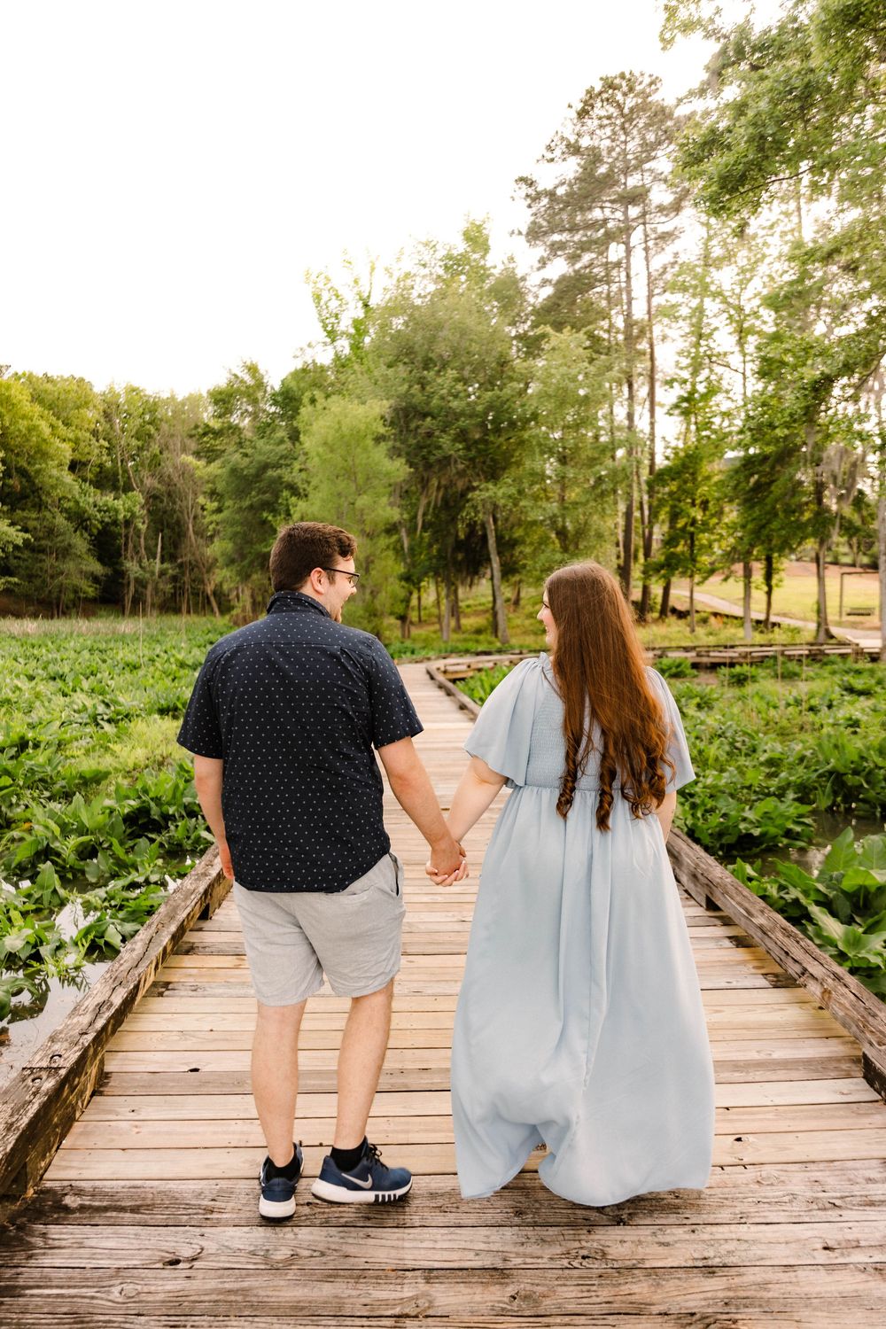 A couple holding hands walks along a wooden boardwalk surrounded by lush greenery and tall trees on a sunny day.
