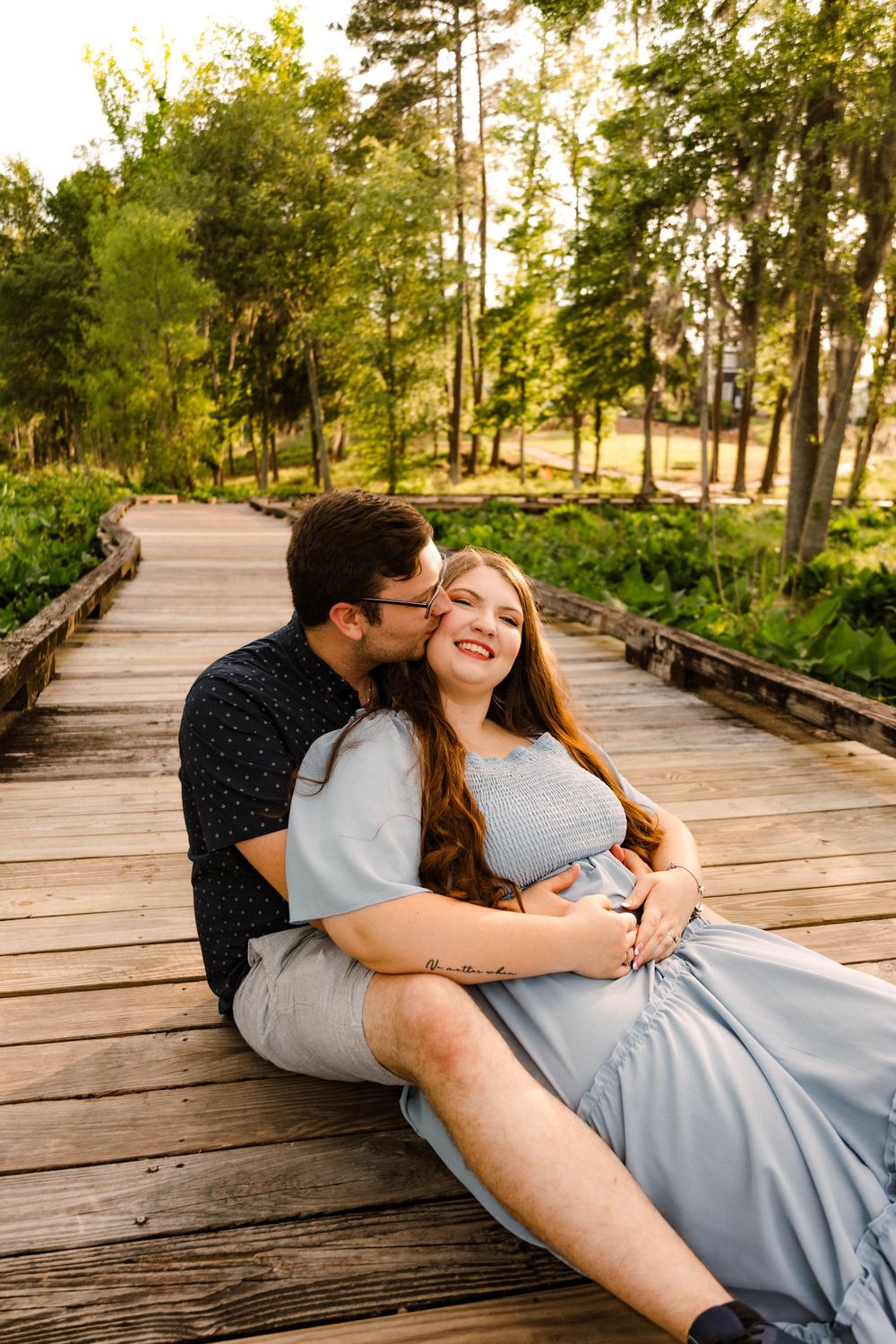 A couple shares a tender moment while sitting on a wooden boardwalk surrounded by trees during golden hour sunset.