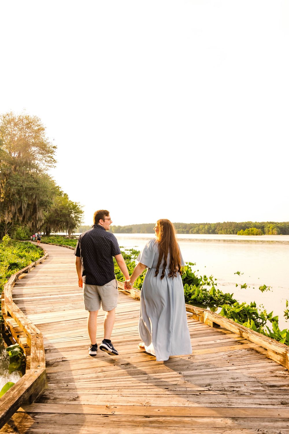 A couple walks hand in hand on a wooden boardwalk along a scenic waterfront during golden hour.