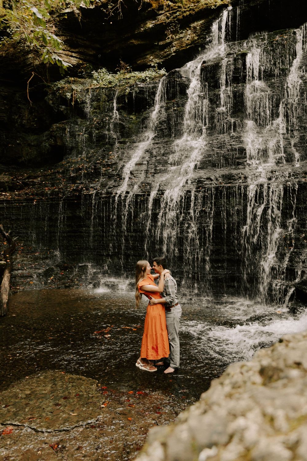 A couple embraces in front of a cascading waterfall with autumn leaves and rocks surrounding them.