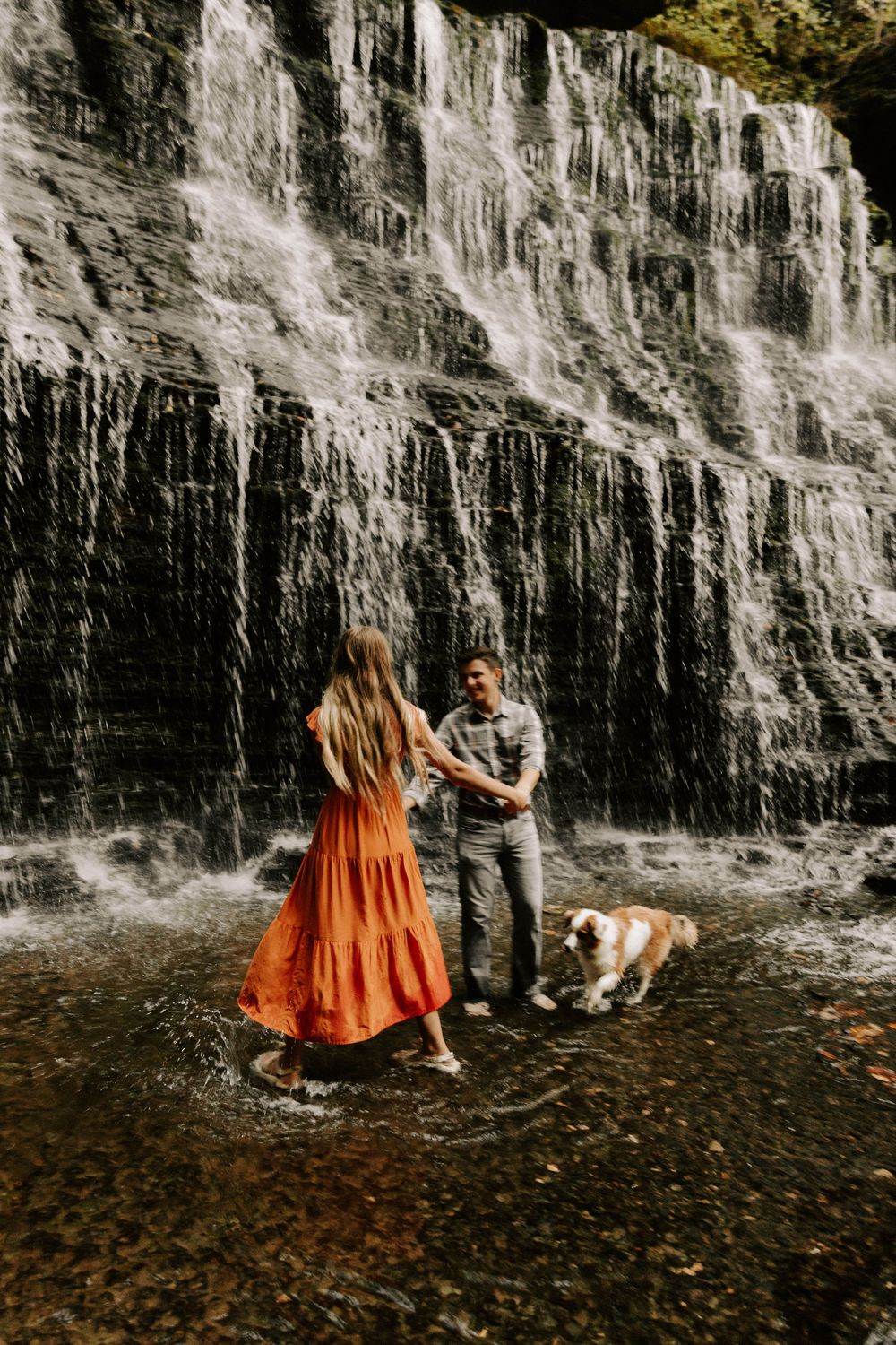 A couple and their dog stand in the shallow water beneath a majestic cascading waterfall on an autumn day.
