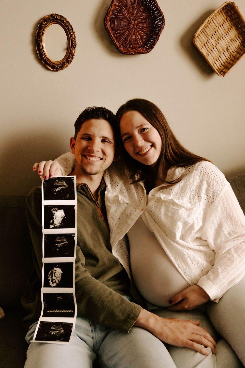 A couple sits together on a couch displaying ultrasound photos while decorative baskets hang on the wall behind them.
