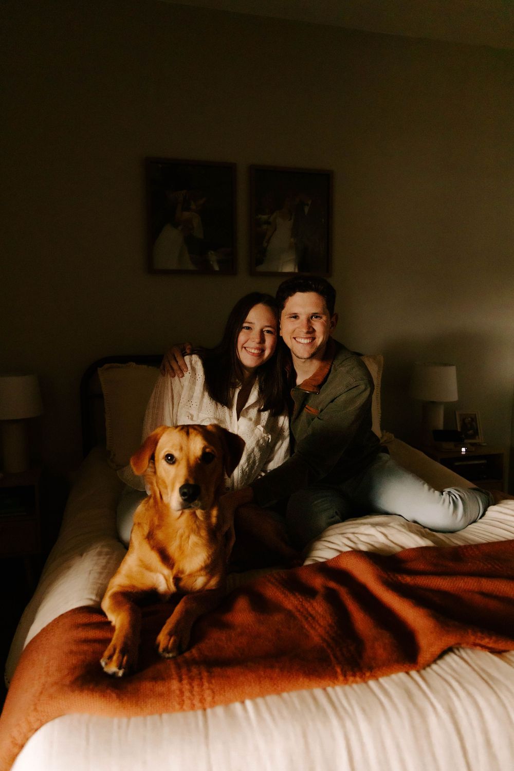 A golden retriever dog rests on a red blanket on a bed with two people in a warmly lit bedroom interior.