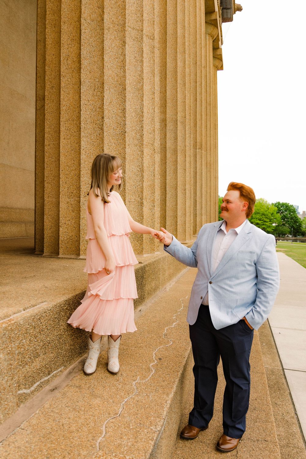 Two people standing by classic stone columns in formal attire share a tender moment at the Parthenon replica.