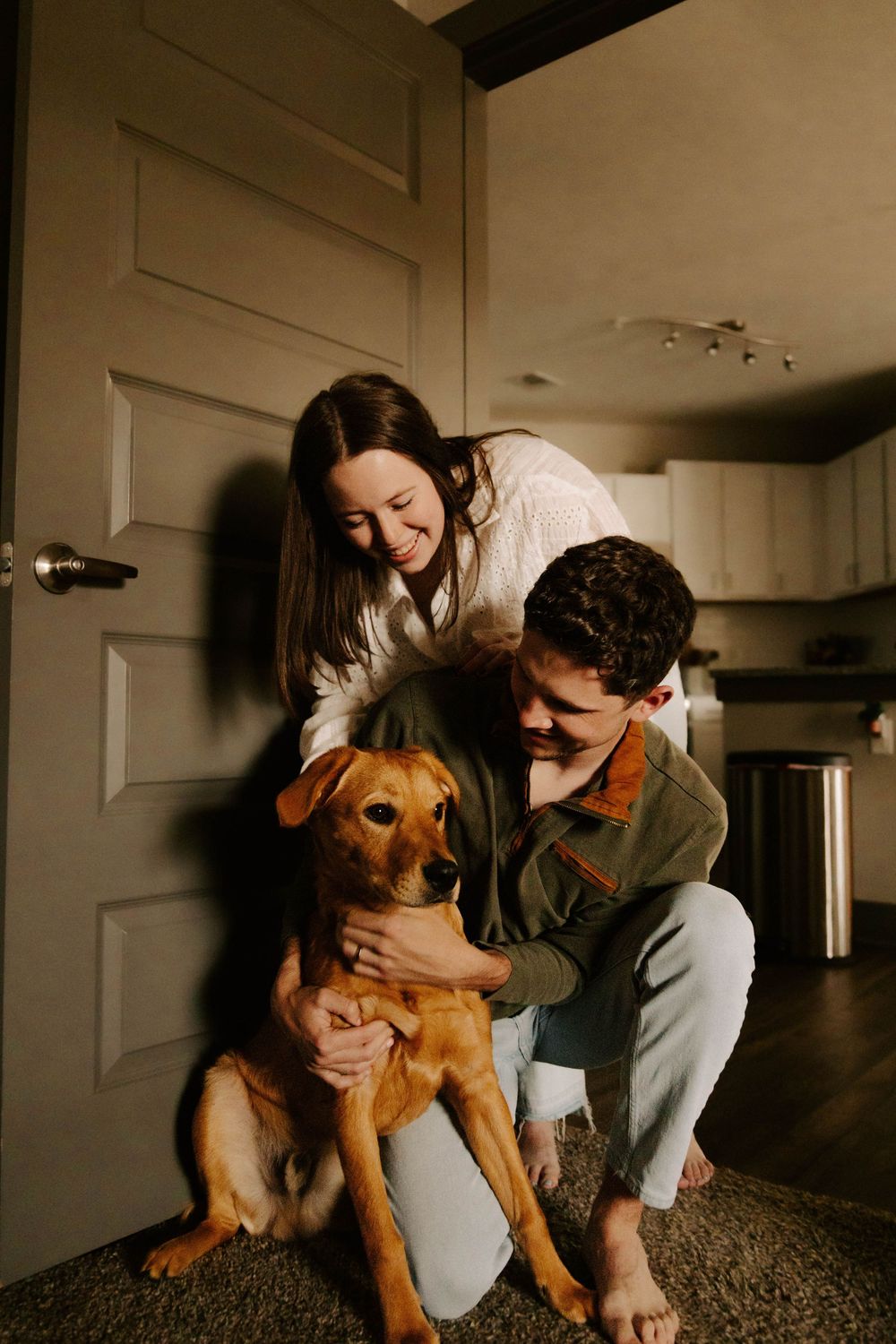A couple sitting on carpet lovingly petting their golden retriever mix dog in a cozy interior setting.