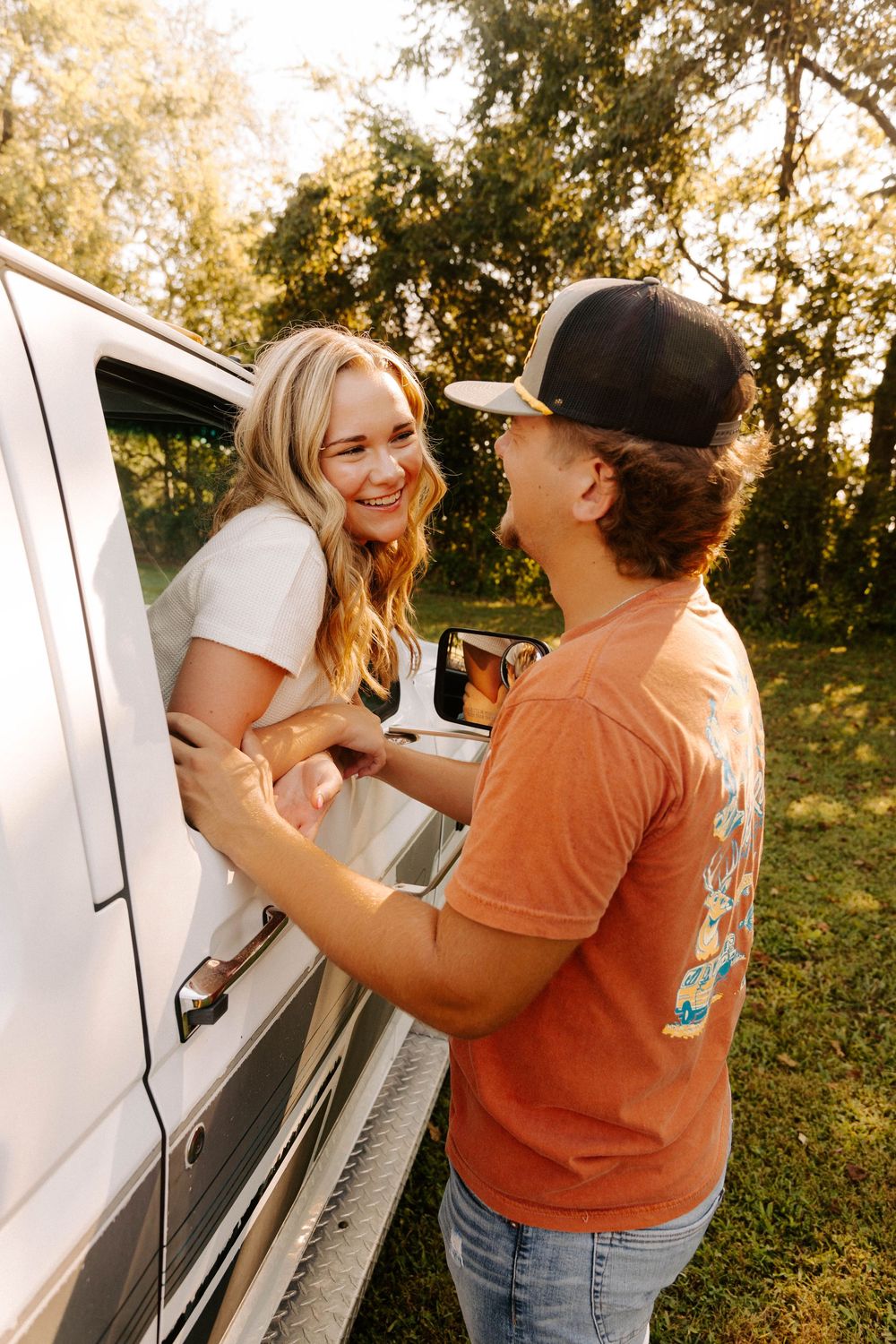 Two young people sharing a joyful moment beside a white vehicle in warm golden sunlight outdoors.