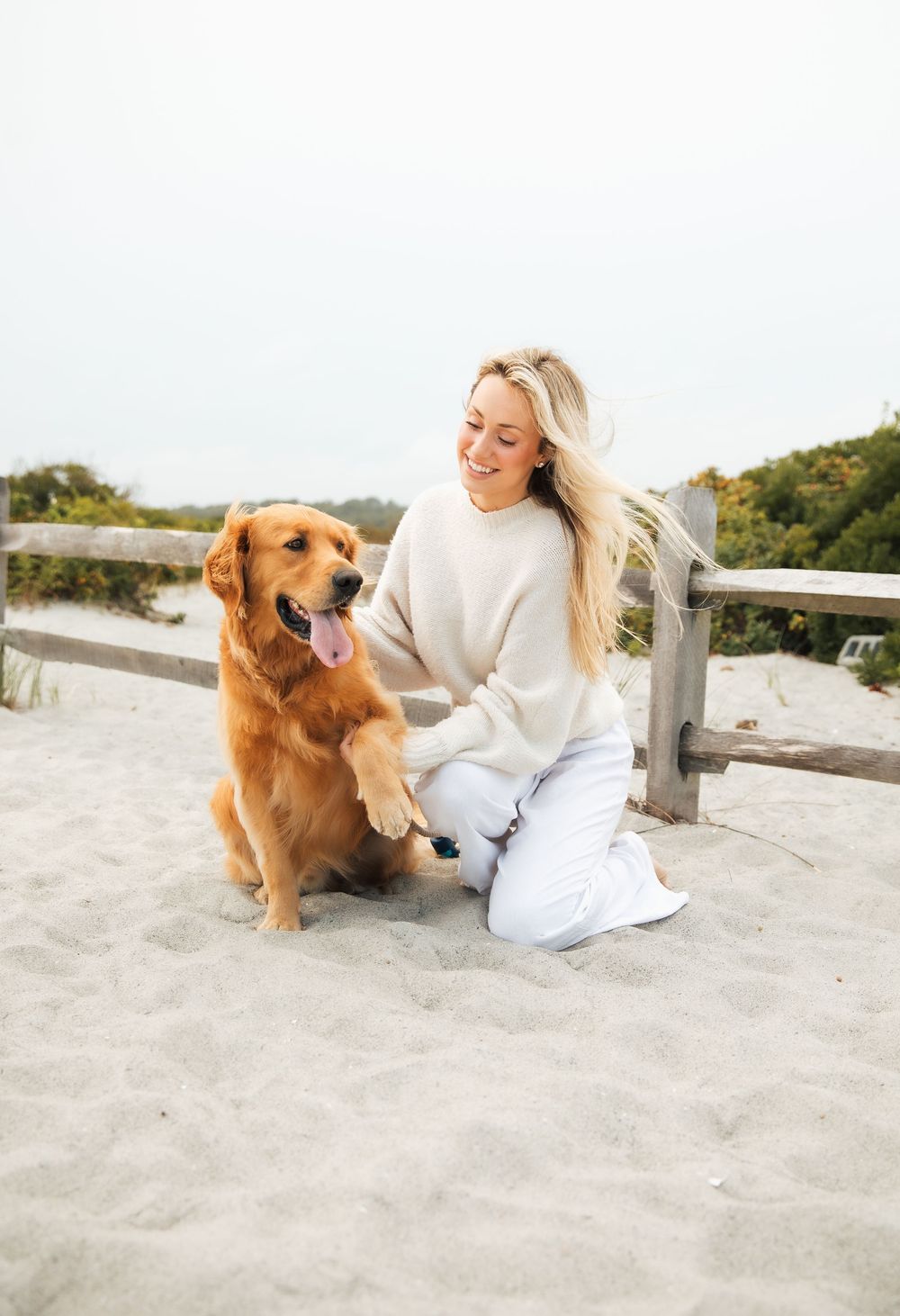 Smiling blonde woman in cream sweater sitting on sandy beach with golden retriever dog raising paw. Third Beach Middletown Rhode Island