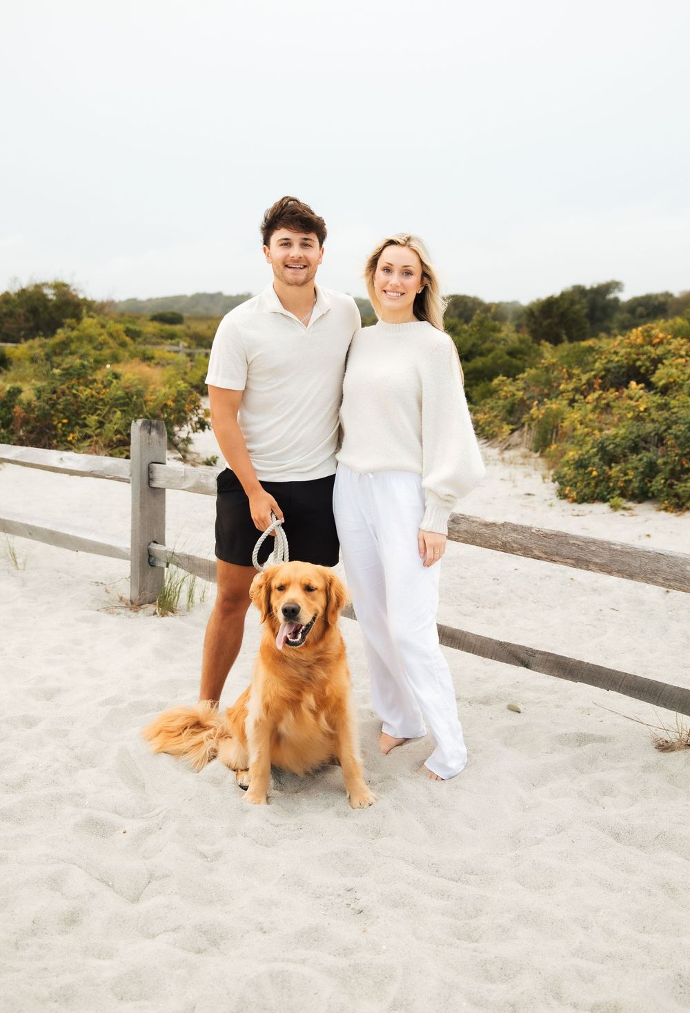 A smiling couple in white tops poses with a golden retriever on a sandy beach boardwalk. Third Beach Middletown Rhode Island