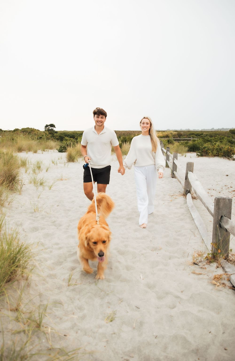 A couple walks a golden retriever on a sandy beach path, holding hands on a bright, breezy day. Third Beach Middletown Rhode Island