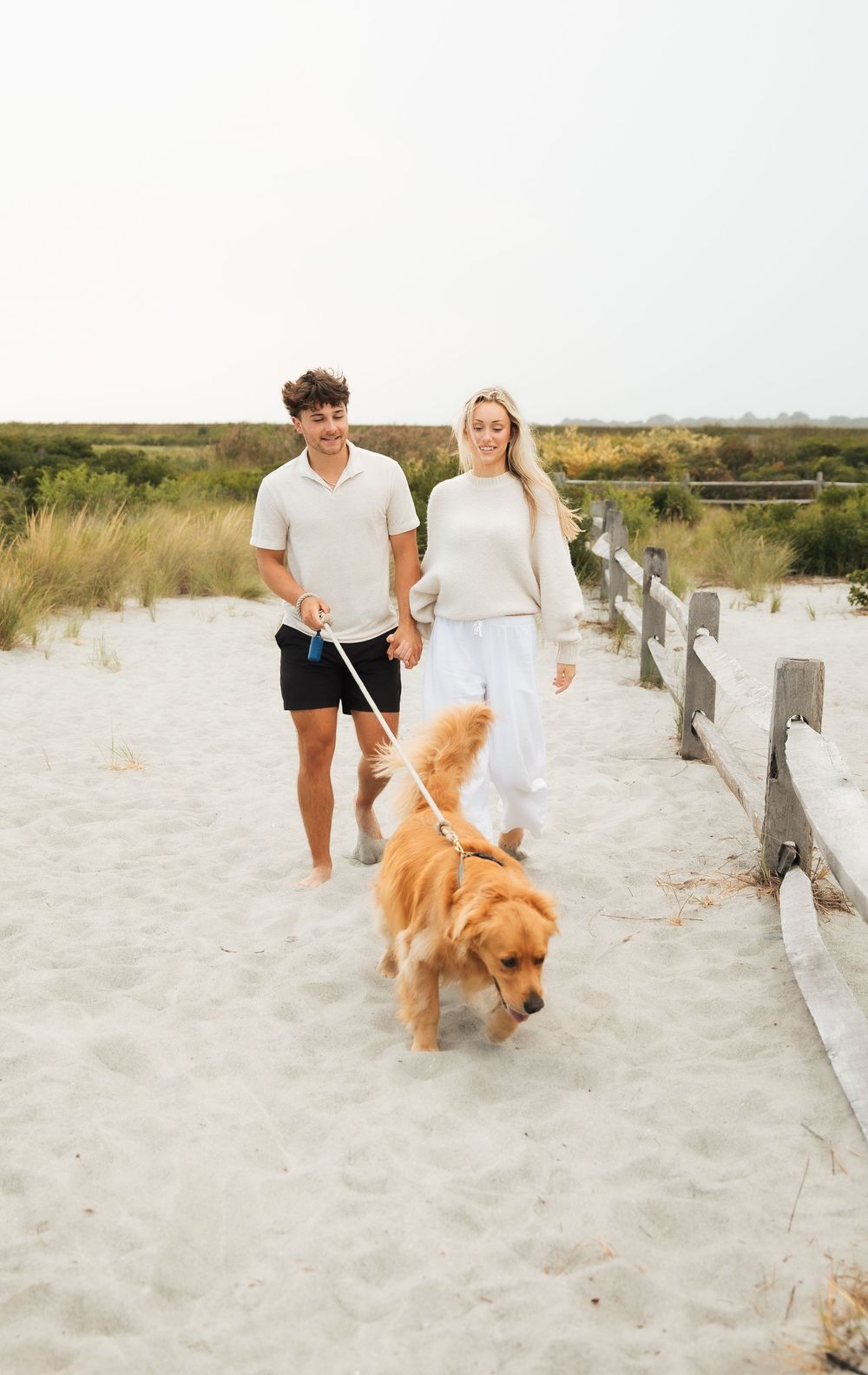 A smiling couple walks a golden retriever on a white sandy beach path with dunes and wooden fence. Third Beach Middletown Rhode Island