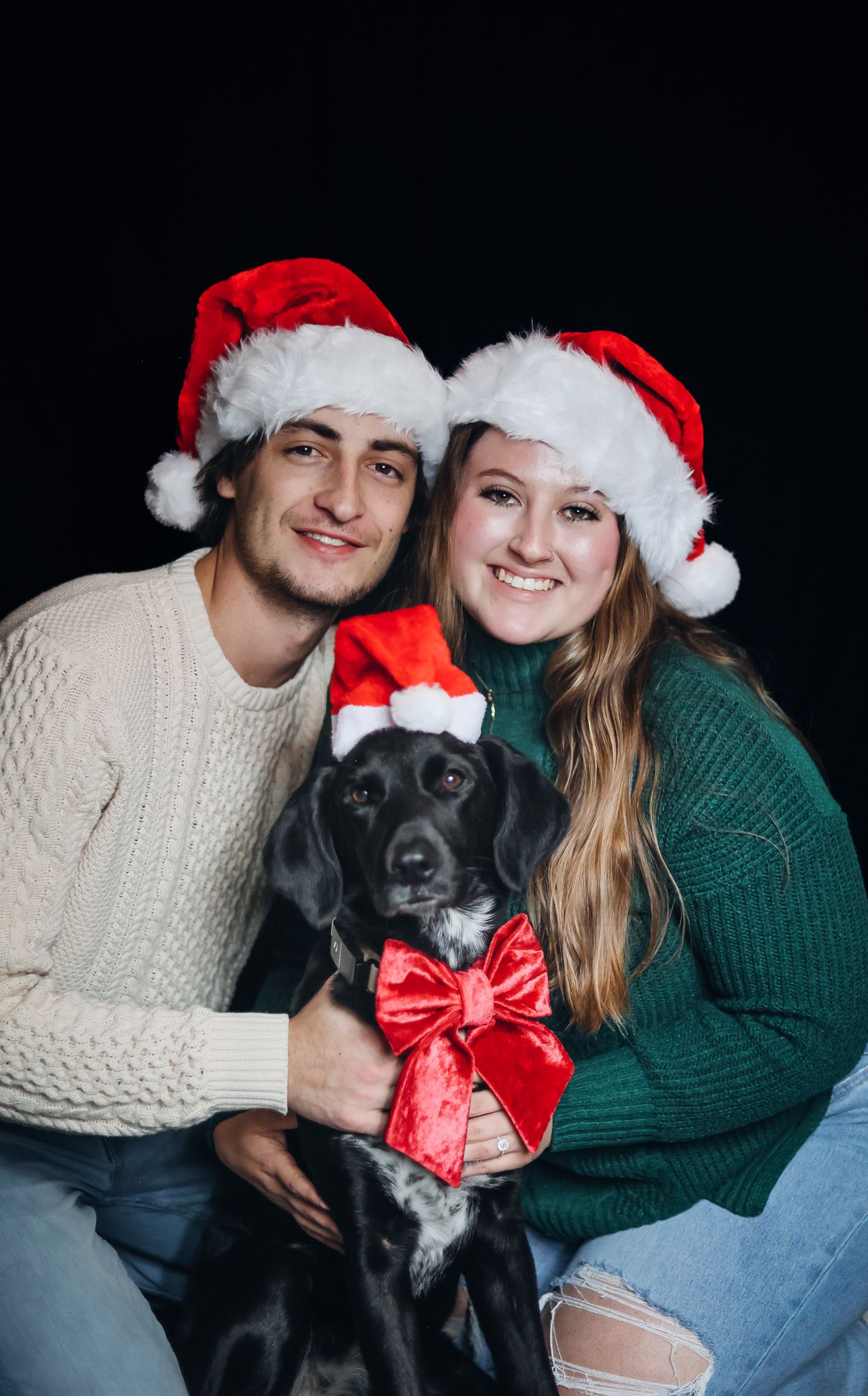 Holiday portrait with black dog wearing red bow between two people in Christmas hats.family Christmas session
