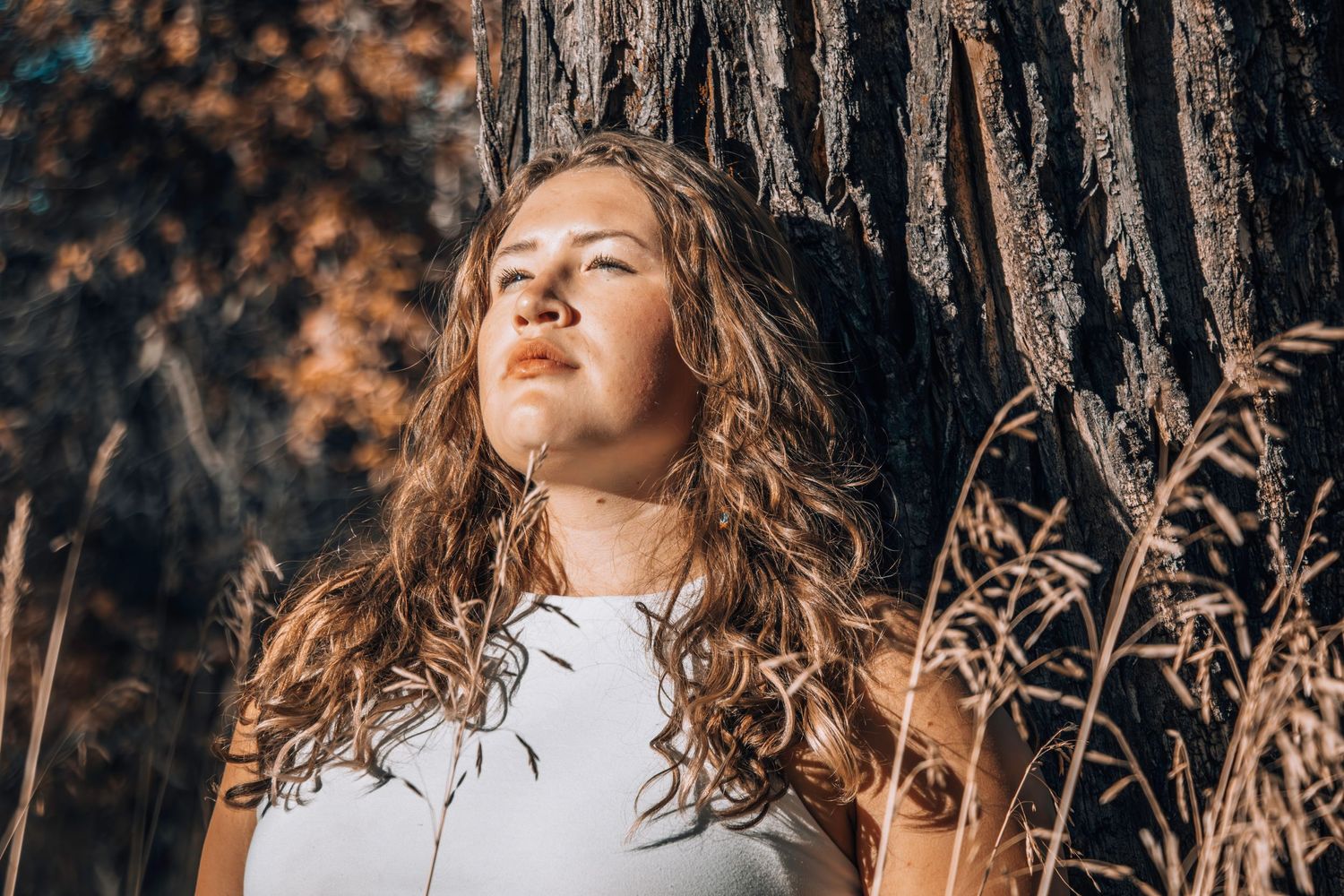 Person with long wavy hair rests against tree trunk in warm autumn sunlight.
