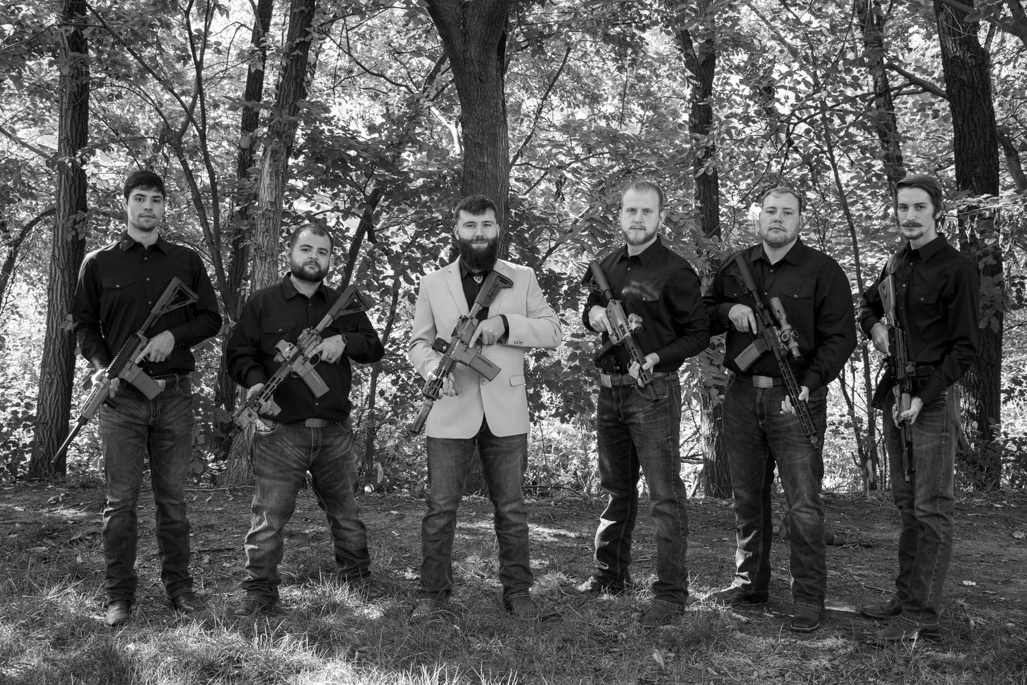 Wedding party members in dark attire pose for black and white photo in wooded setting.