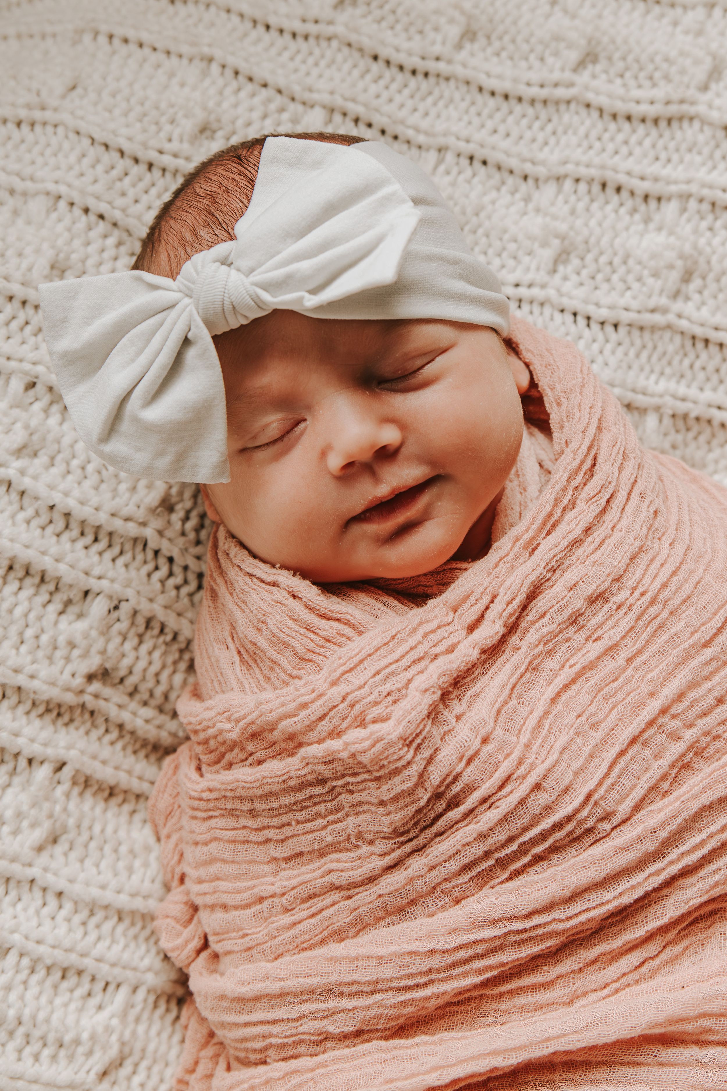 Close-up of peaceful newborn swaddled in coral muslin with white bow headband.