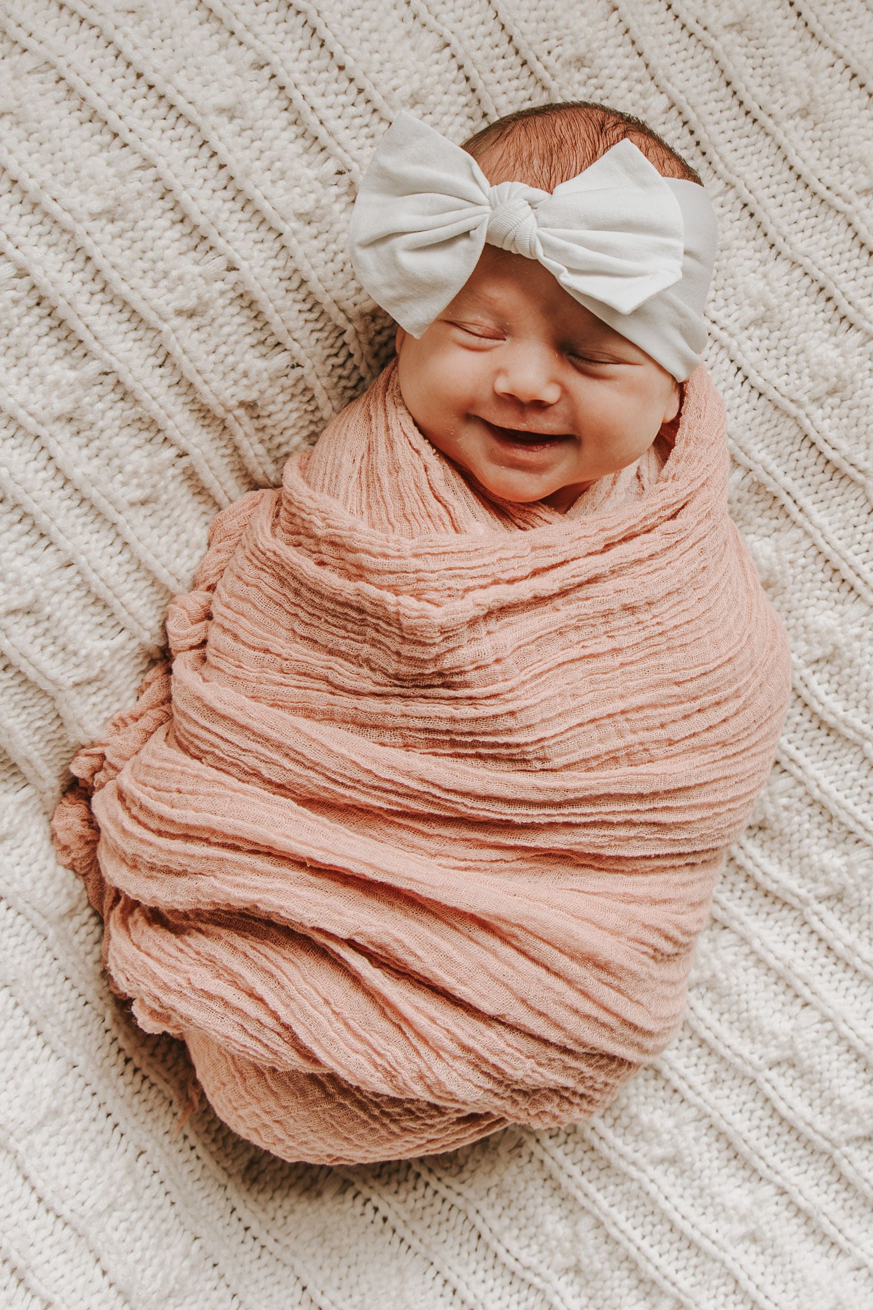 Sleeping newborn wrapped in peach muslin fabric on cream knit blanket with white bow.