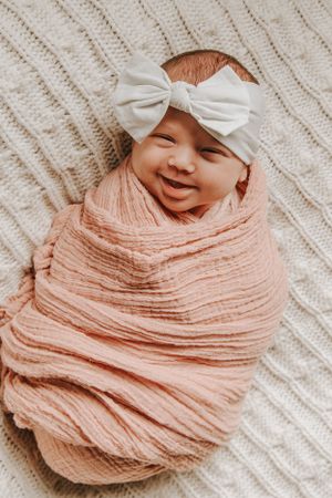 Sweet newborn baby smiling while wrapped in peach muslin on cream knit background.