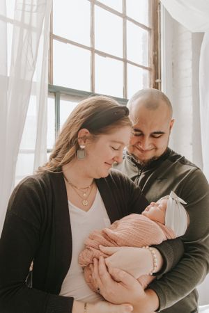 Family sharing tender moment with newborn by sunlit windows with flowing white curtains.