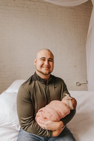 A couple cradles their sleeping newborn baby wrapped in a pink swaddle against a white brick wall.