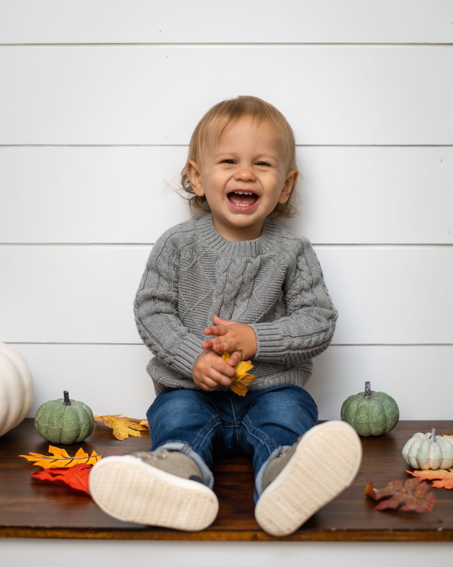 A toddler in a gray sweater sits with fall decorations and pumpkins.