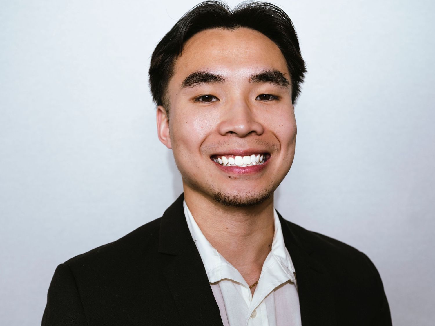 Young man in black blazer and white shirt smiling confidently against a light gray background.