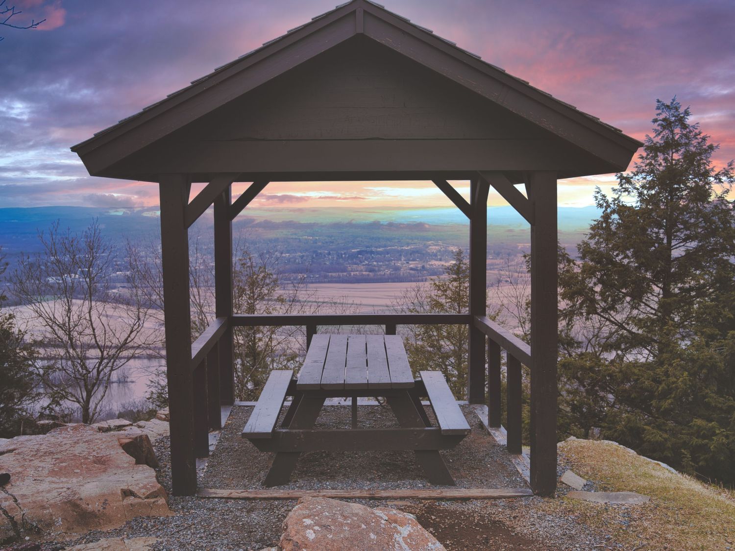 Rustic wooden gazebo with picnic table overlooking breathtaking mountain valley at colorful sunset.