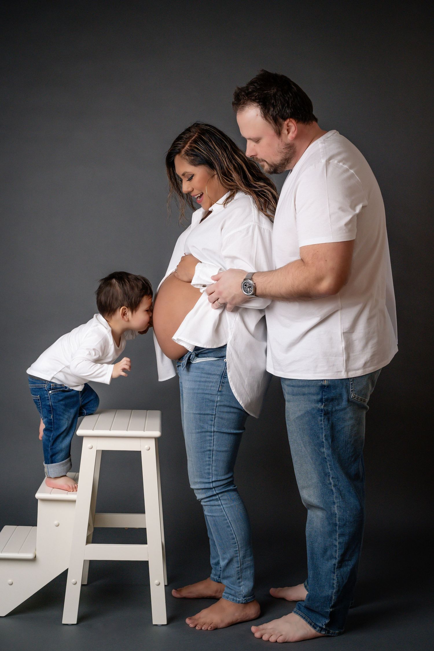 Family in white shirts and jeans interacting with small child on white stool against dark background.